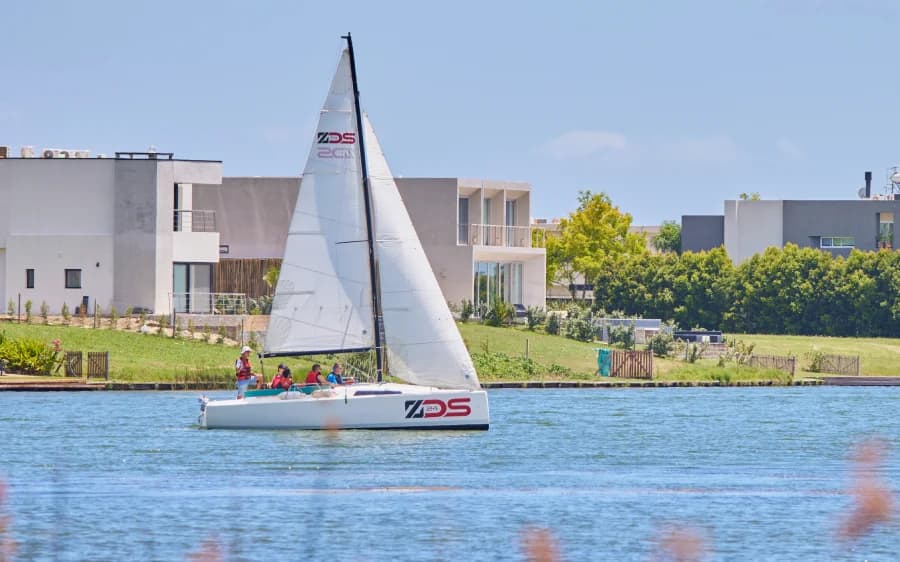Velero navegando en la laguna de Puertos, con casas y juncos en la orilla