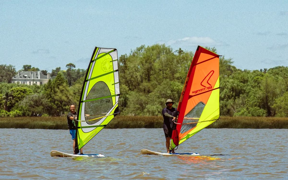 Dos personas navegan en windsurf, con una vela amarilla y otra roja, frente a la costa arbolada