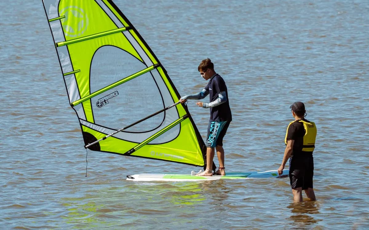 Un instructor, parado en el agua, acompaña a un alumno que levanta la vela de windsurf